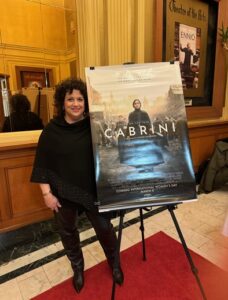New American Integration Director Denise Phillips Beehag smiles with the official Cabrini movie poster shortly before she spoke to the packed crowd of film-goers about IIB's important work community